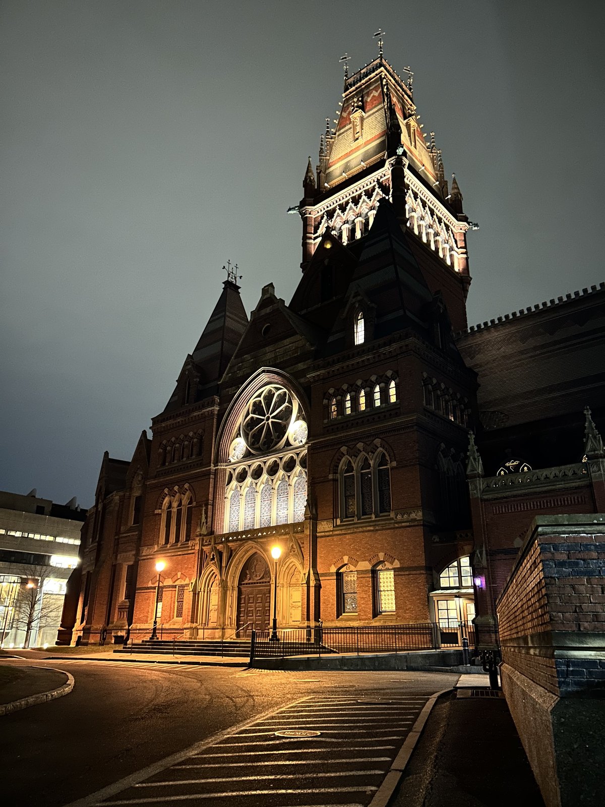 Night view of a red-brick Gothic Revival building with a lit rose window and clock tower, an empty street in front