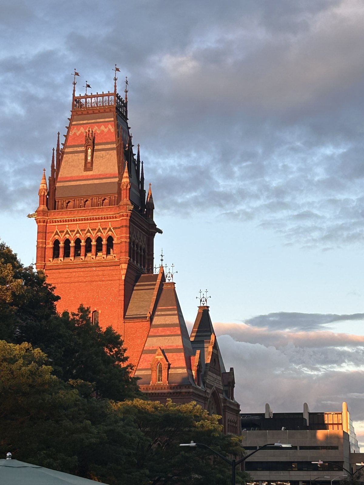 A red-brick Gothic Revival clock tower glowing orange at dusk against a cloudy sky