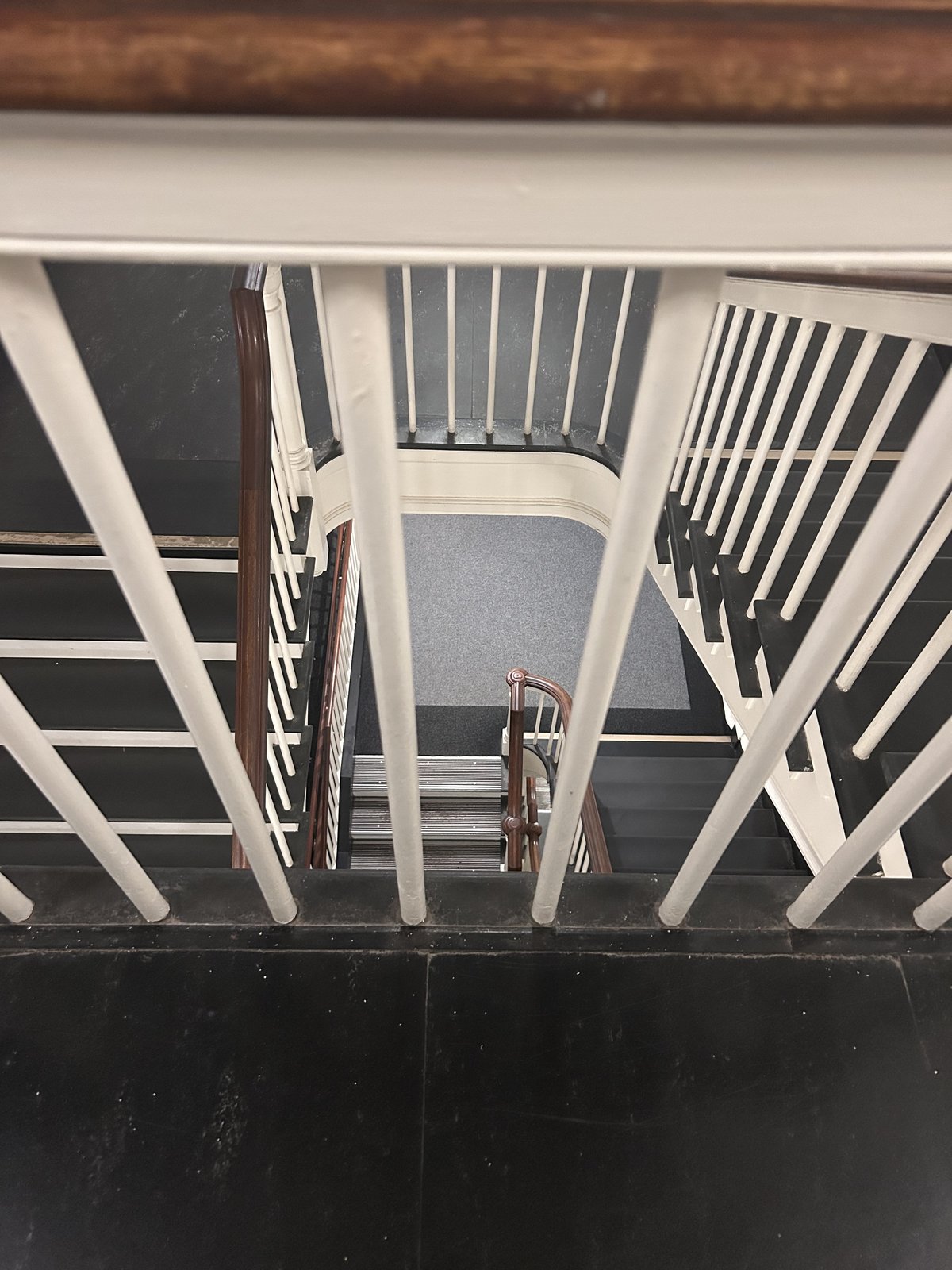 Looking down a white wooden staircase and railing toward the floor below, with a wooden handrail in the foreground